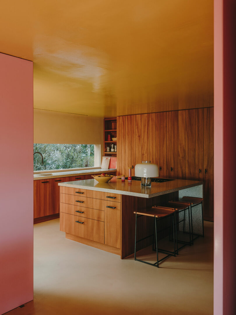 La cocina moderna cuenta con gabinetes de madera, isla con taburetes, paredes y techos de color rosa y amarillo, electrodomésticos y ventanas con vista a la vegetación.