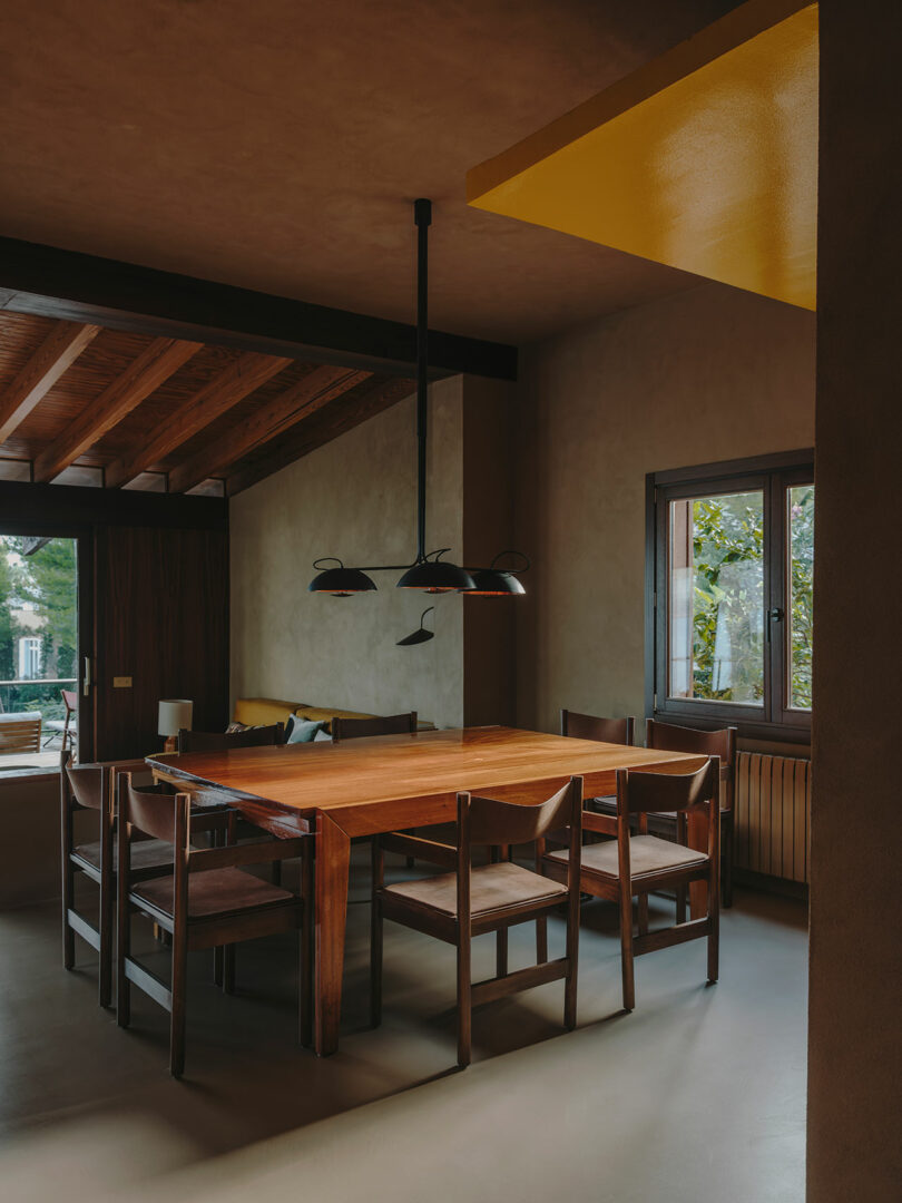 Con vigas de madera, paredes beige y una ventana que da a la vegetación exterior, la mesa de comedor de madera de la habitación y seis sillas a juego se encuentran debajo de una moderna lámpara de araña negra.