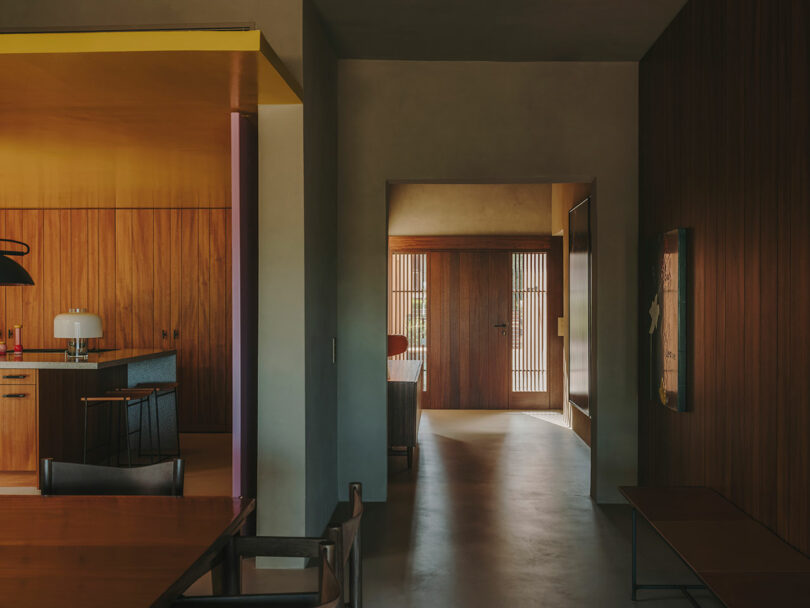 Interior moderno con paredes con paneles de madera, zona de cocina abierta, mesa de comedor y pasillo iluminado que conduce a la puerta principal con listones verticales.