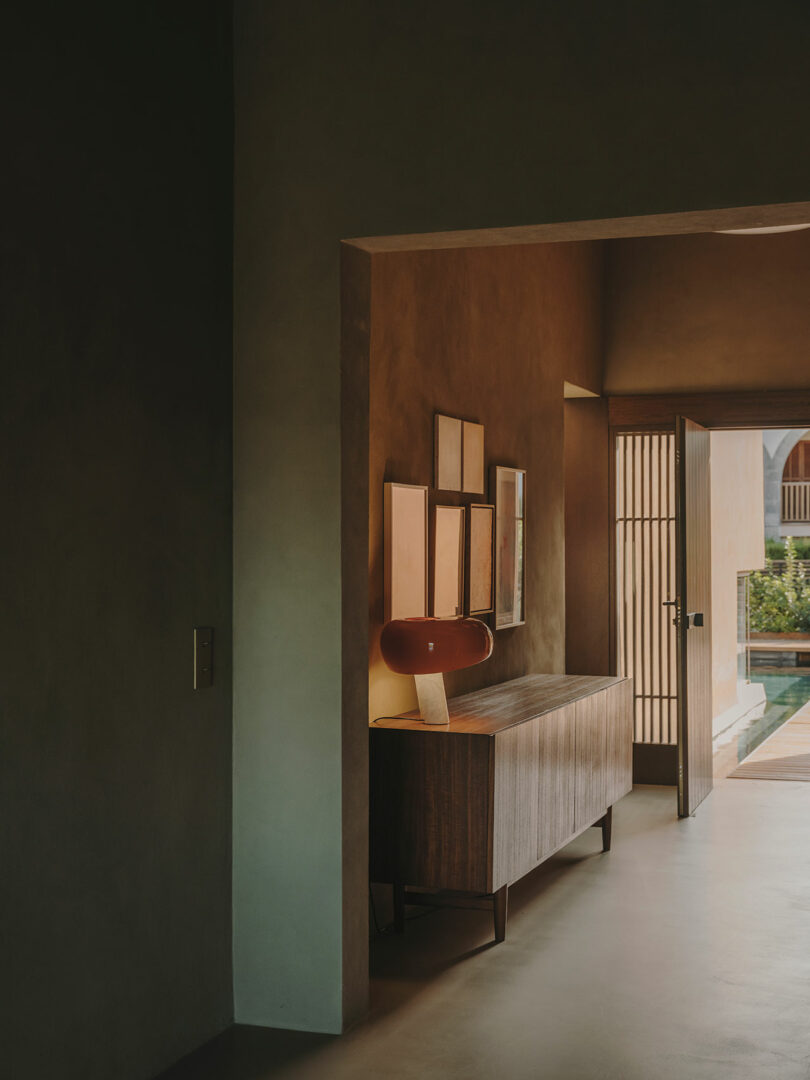 El estrecho pasillo con paredes de colores pastel cuenta con aparadores de madera, luces rojas, cinco cuadros enmarcados y una puerta abierta que conduce a un área exterior soleada y arbolada.