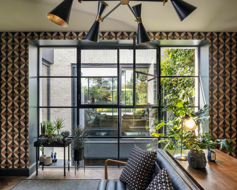 Modern living room with geometric wallpaper, large black-framed window, various indoor plants, a black sofa with patterned cushions, and a desk lamp on a wooden table.