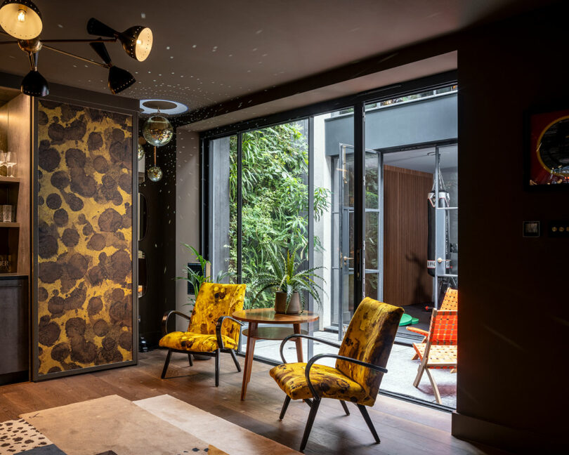 Modern living room with two yellow-patterned chairs, a small round table, and glass doors opening to a patio with orange chairs and lush greenery outside.