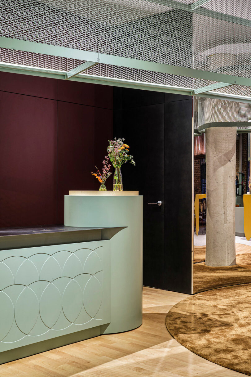A modern interior at NOXX Hotel features a mint green reception desk, vases with flowers, a cylindrical concrete column, and geometric ceiling panels over wood and carpet flooring.