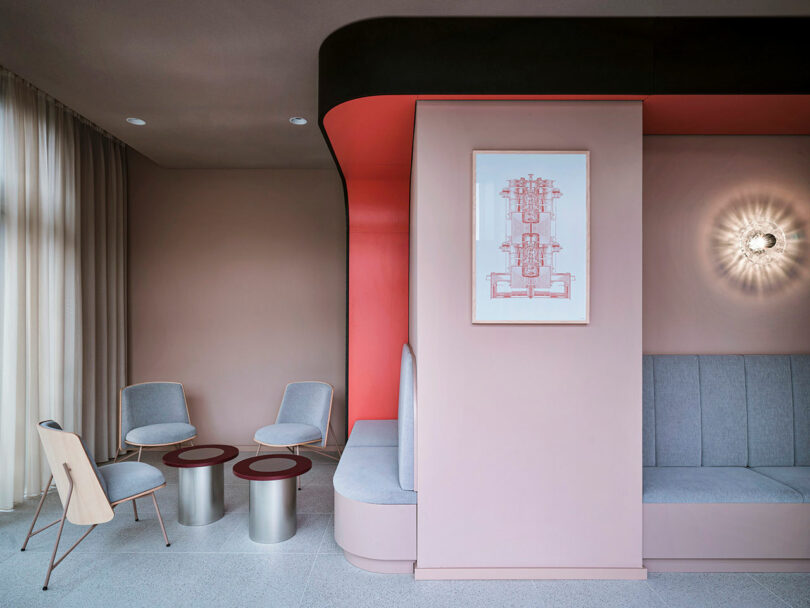 Modern lounge at NOXX Hotel with pastel pink and gray seating, round tables, wall art, and a contemporary sconce; accented by a bold red curved architectural feature.