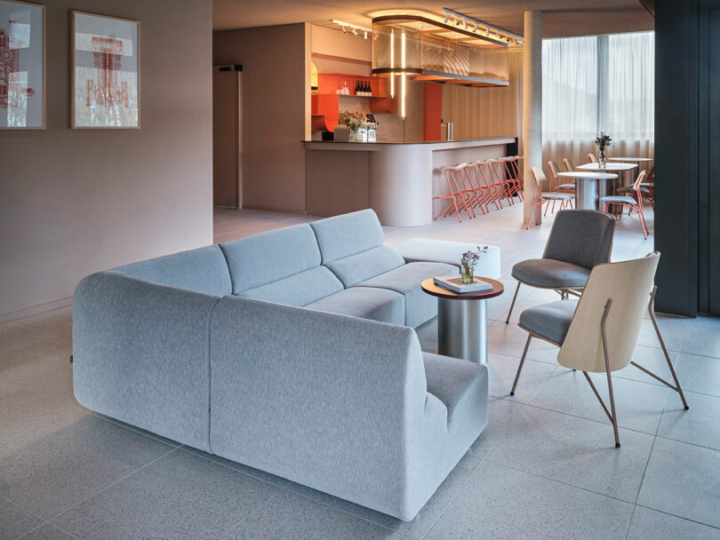 Modern lounge area at NOXX Hotel featuring a light gray sectional sofa, two accent chairs, small tables, and a bar with stools in the background under warm lighting.