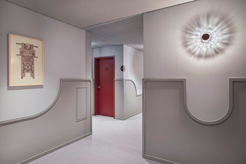 A hallway at NOXX Hotel with light gray walls, a red door, a round wall lamp emitting soft light, and framed artwork decorating one wall.