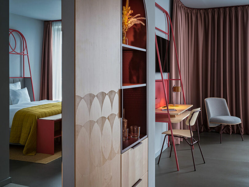 Modern NOXX Hotel room with a double bed, yellow blanket, desk, chair, and pink accents, divided by a wooden partition featuring built-in shelves and geometric patterns.