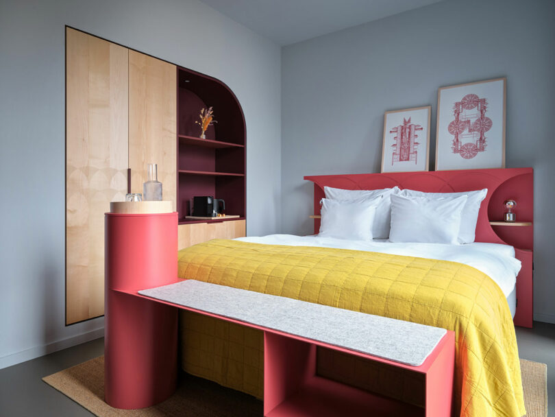 Modern bedroom at NOXX Hotel with a red headboard, yellow quilt, wooden accents, built-in shelving, and framed artwork above the bed against a light gray wall.