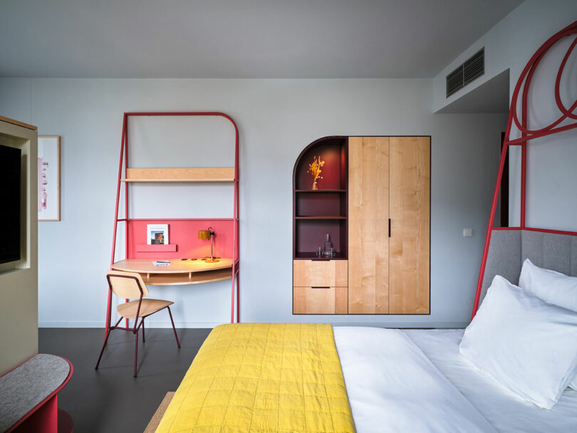Modern NOXX Hotel room featuring a neatly made bed with a yellow blanket, built-in wooden wardrobe, and a red desk with chair set against a light gray wall.
