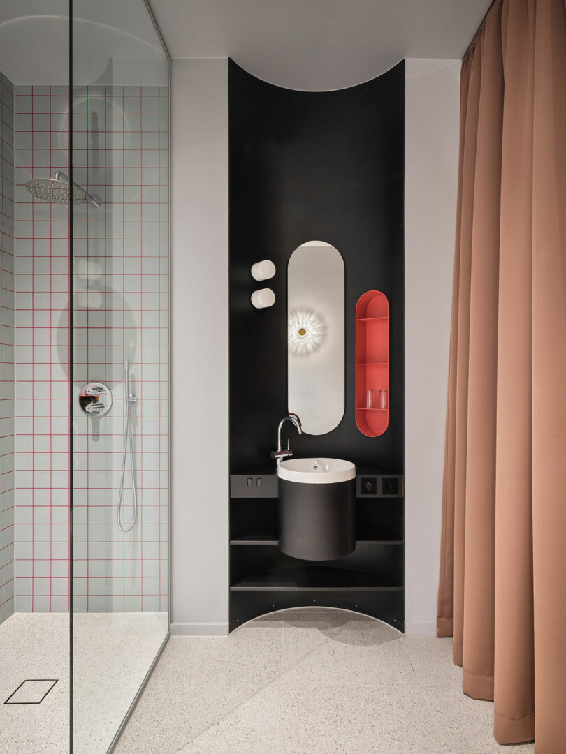 Modern bathroom at NOXX Hotel with a glass shower on the left, a black vanity featuring a round sink and oval mirror center, red shelving, and a beige curtain on the right.