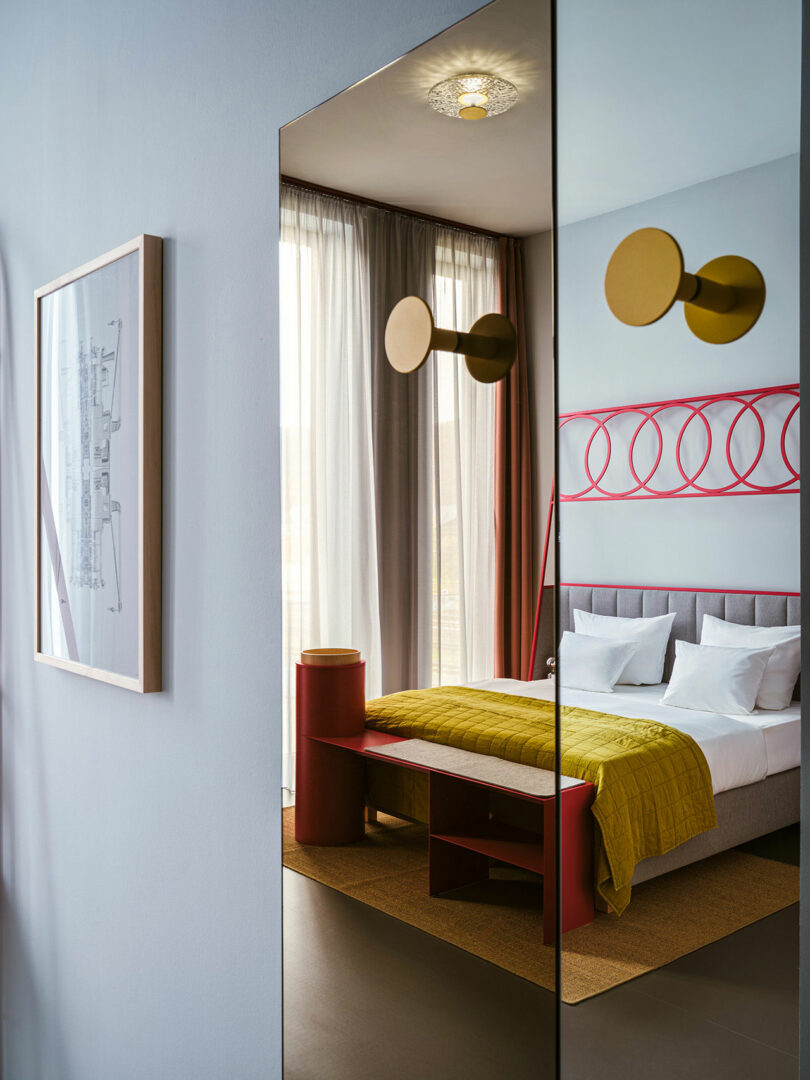 A modern NOXX Hotel bedroom with a gray bed, yellow blanket, white pillows, red frame, large window with sheer curtains, and a mirror reflecting the room.