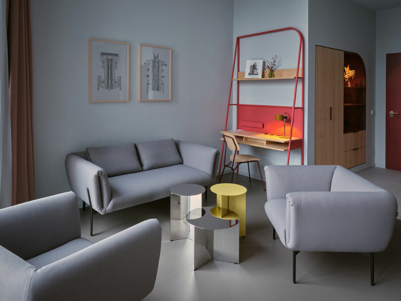 Modern living room at NOXX Hotel with gray sofas, metallic and yellow side tables, stylish wall art, and a red-framed desk with a wooden shelf set against a sleek gray wall.