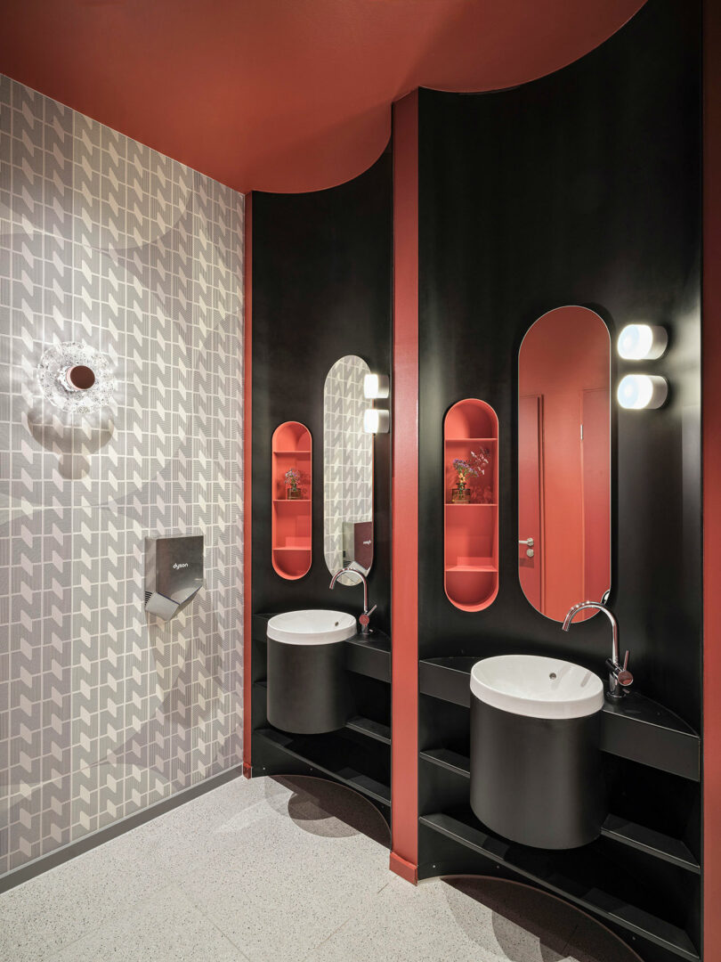 Modern NOXX Hotel bathroom with a patterned tile wall, two round sinks in black and red cabinetry, oval mirrors, wall lights, and built-in shelves.