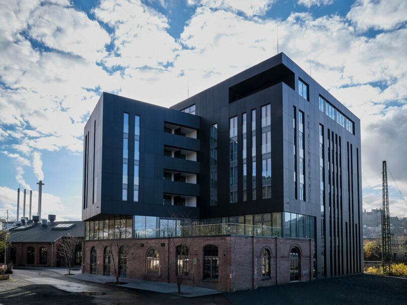 A modern black building with large glass windows, now home to the NOXX Hotel, is built on top of an old brick industrial structure under a partly cloudy sky.