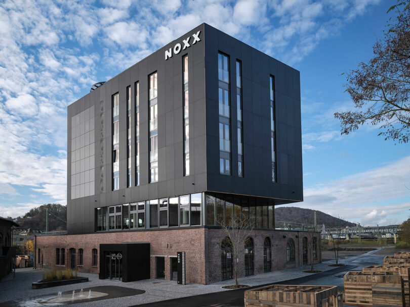 A modern building with a brick lower level and a black upper section labeled "NOXX Hotel," set against a partly cloudy sky with surrounding paved areas.