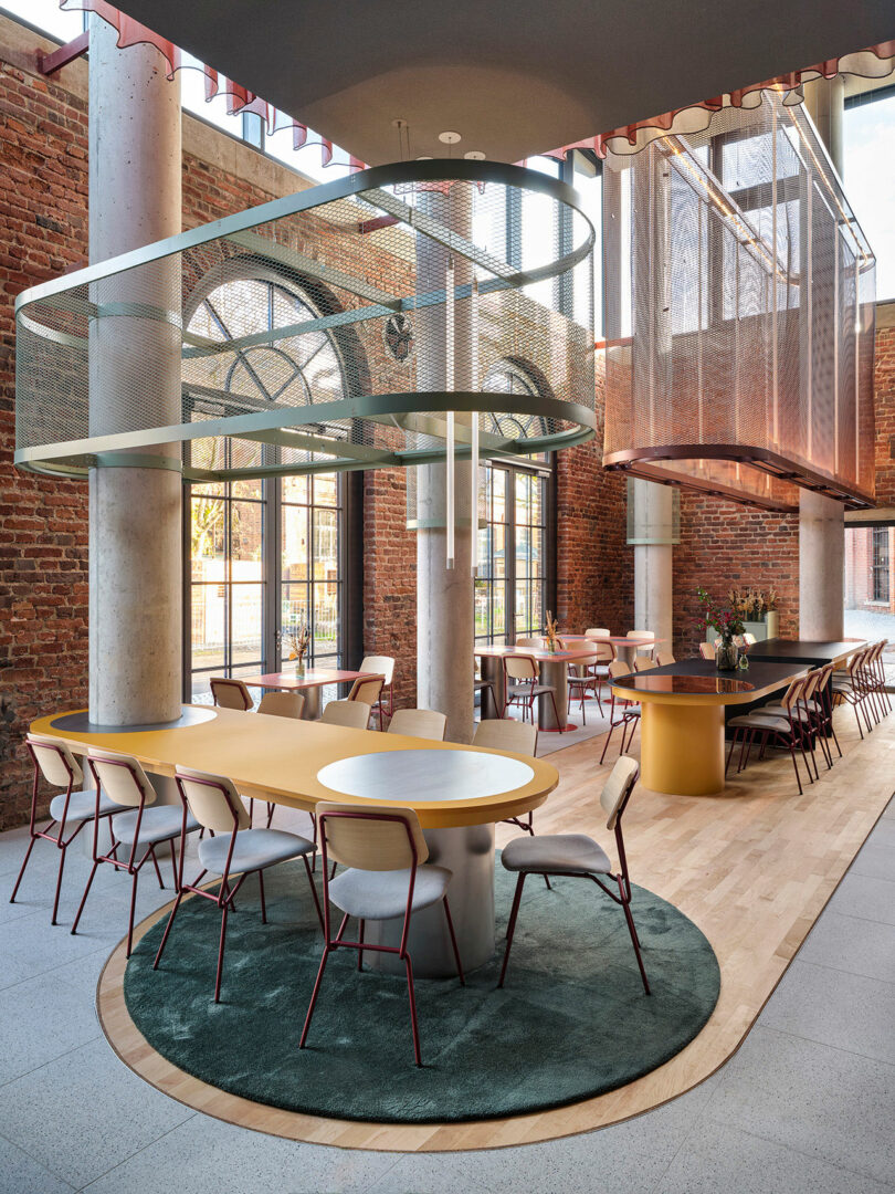 Modern communal dining area at NOXX Hotel with round and rectangular tables, contemporary chairs, exposed brick walls, large windows, and decorative metal mesh structures suspended from the ceiling.