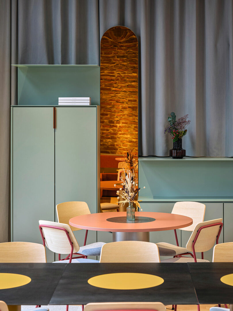 A modern room at NOXX Hotel features pastel green cabinets, a round coral table, light chairs, a vase with dried flowers, and a curtained doorway revealing a softly lit brick wall.