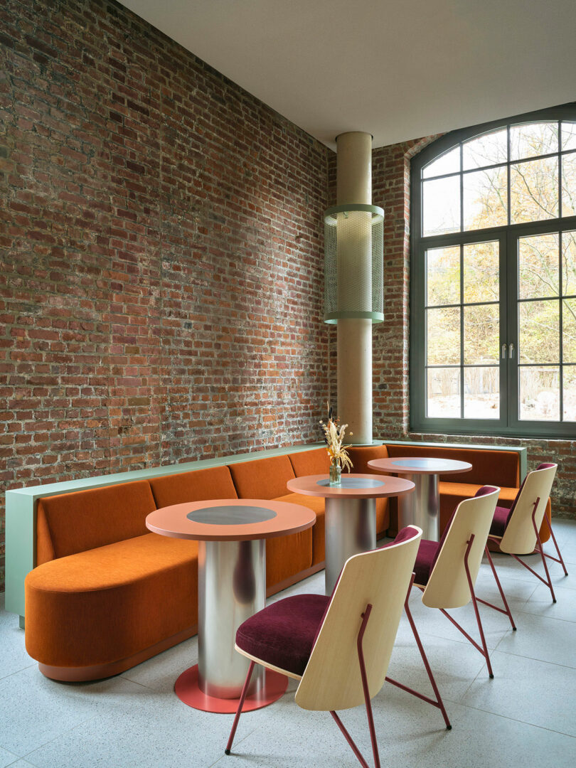 Modern lounge at NOXX Hotel with exposed brick walls, large windows, an orange upholstered sofa, round tables with metal bases, and wooden chairs with burgundy seats.