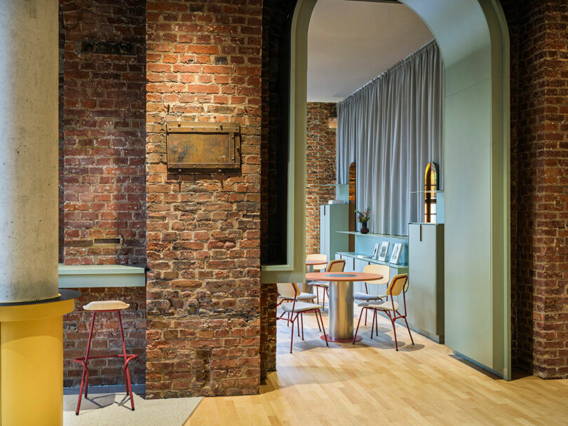 Modern interior at NOXX Hotel featuring exposed brick walls, an archway, light wood floors, a round table with chairs, high stools, and grey curtains in the background.