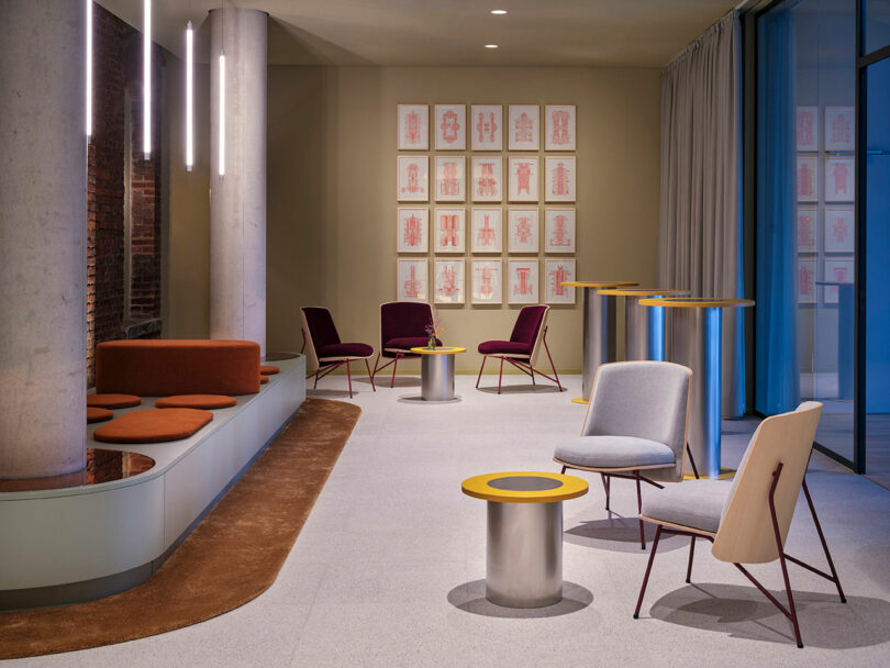 Modern lounge area at NOXX Hotel with contemporary chairs, round tables, a bench with rust-colored cushions, and framed abstract art on the wall. Vertical lights hang stylishly from the ceiling.