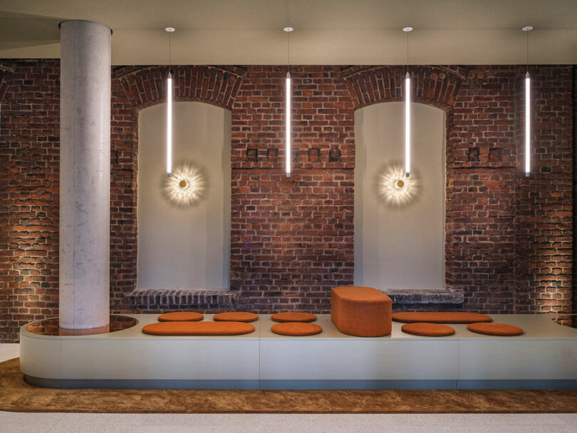 Modern lounge area at NOXX Hotel with a long bench, round orange cushions, and a curved backrest, set against an exposed brick wall featuring decorative lighting and two elegant arched alcoves.