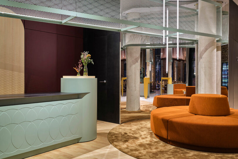 Modern lobby at NOXX Hotel featuring curved orange seating, a mint green reception desk, and exposed concrete columns on light wood flooring. Dried flowers in a vase add a welcoming touch to the counter.