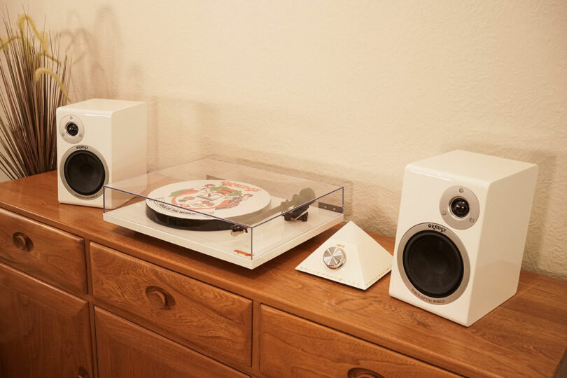 A white turntable with a clear dust cover and a colorful vinyl record sits on a wooden cabinet, flanked by two white speakers and a triangular volume control.