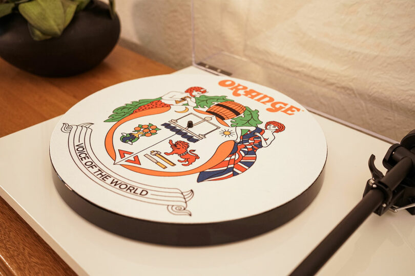 A record player with a decorative slipmat featuring the Orange logo, crest, slogan "Voice of the World," and various colorful illustrations.