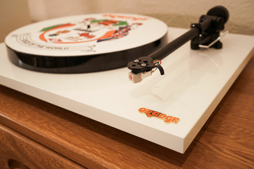 A close-up of a white turntable with a custom illustrated platter and an "Orange" logo on the corner, placed on a wooden surface.