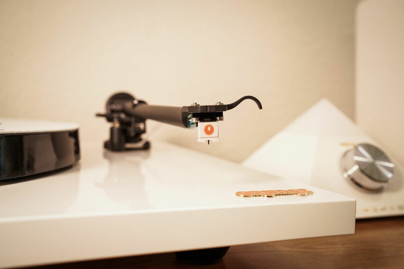 Close-up of a white turntable’s tonearm and cartridge, with a vinyl record partially visible on the left and an audio device with a large knob in the background.
