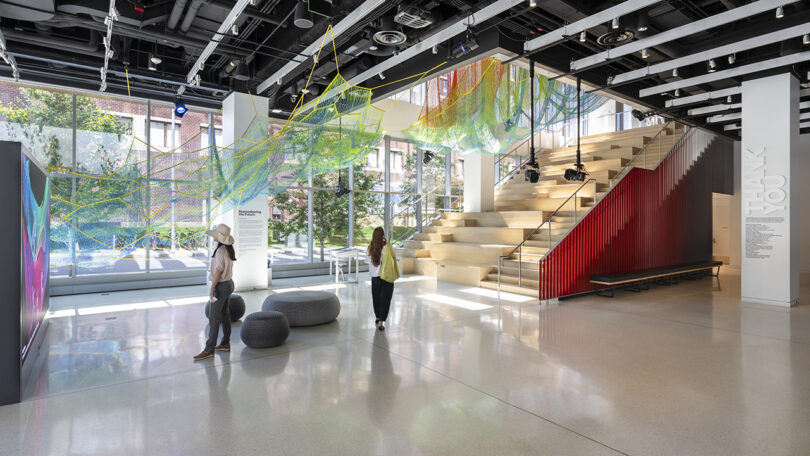 A modern, sunlit interior space with large windows, abstract ceiling art, a wide staircase with red railings, and two people observing the surroundings.