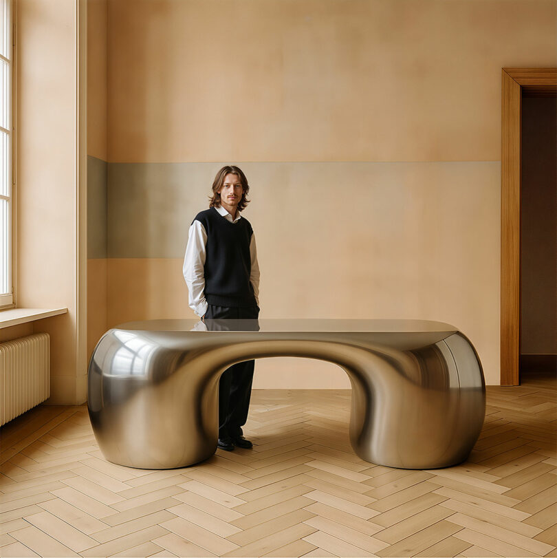 Paddy Pike stands in a minimalist room, dressed in a black vest and white shirt, beside a large, curved, metallic table with a hollow center.