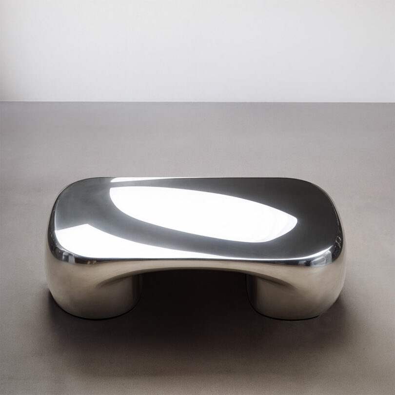 A polished, metallic Paddy Pike rectangular coffee table with rounded edges and a seamless, modern design sits on a plain, gray floor against a white wall.