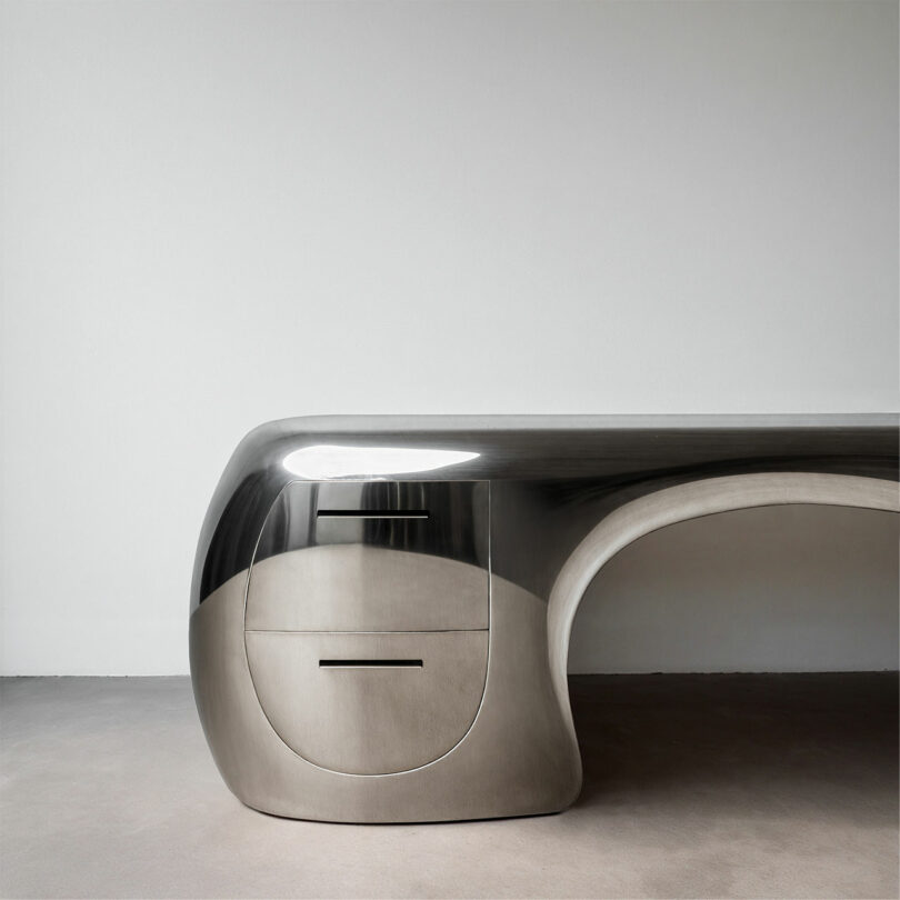 Sleek, metallic, modern Paddy Pike desk with curved edges and built-in drawers sits elegantly against a plain white wall and light floor.