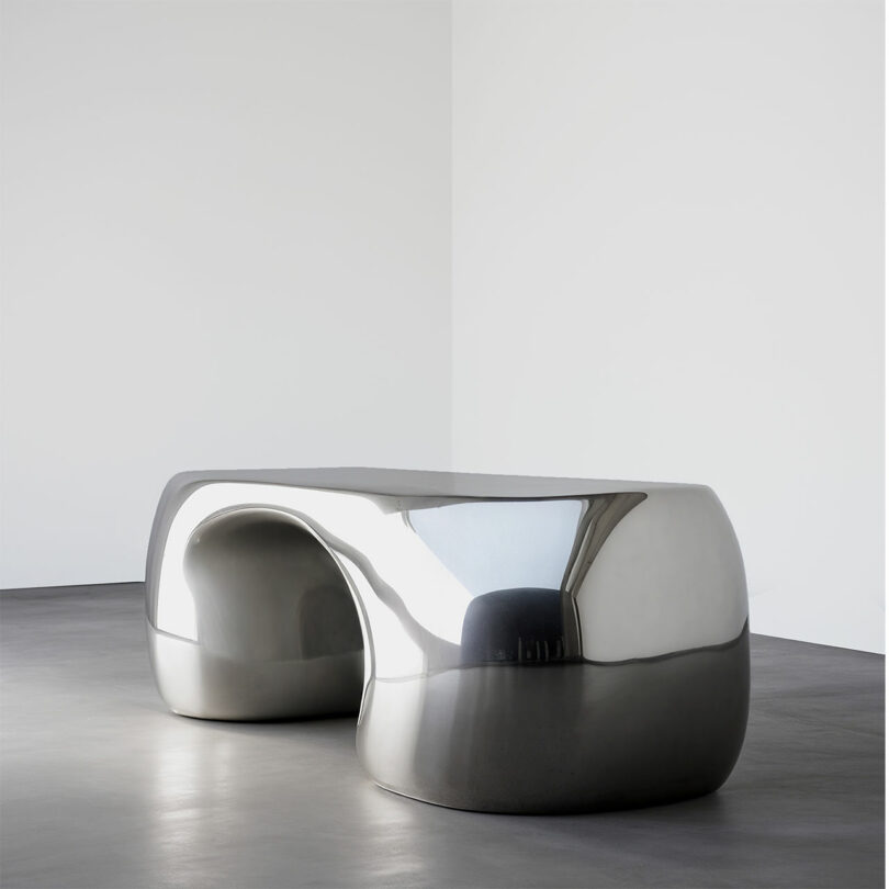A smooth, reflective, metallic bench with rounded edges by Paddy Pike sits in an empty, minimalist room with gray flooring and white walls.