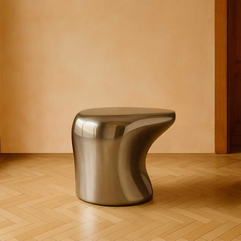 A smooth, metallic, sculptural stool by Paddy Pike with organic curves is placed on a wooden herringbone floor against a beige wall indoors.