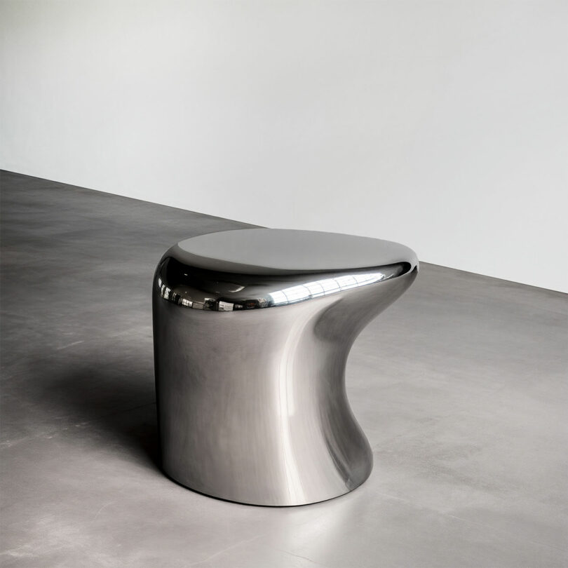 A smooth, metallic, abstract-shaped Paddy Pike stool sits on a polished concrete floor against a plain white wall.