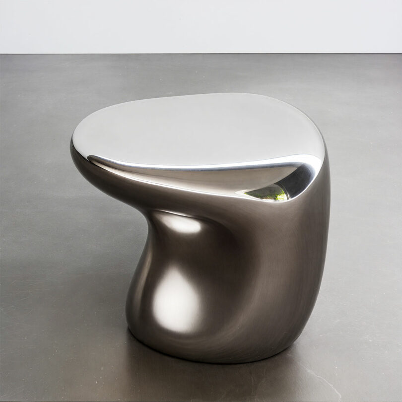 A smooth, metallic, asymmetrical table or stool by Paddy Pike features a reflective surface and organic, curved shape, beautifully placed on a gray floor.