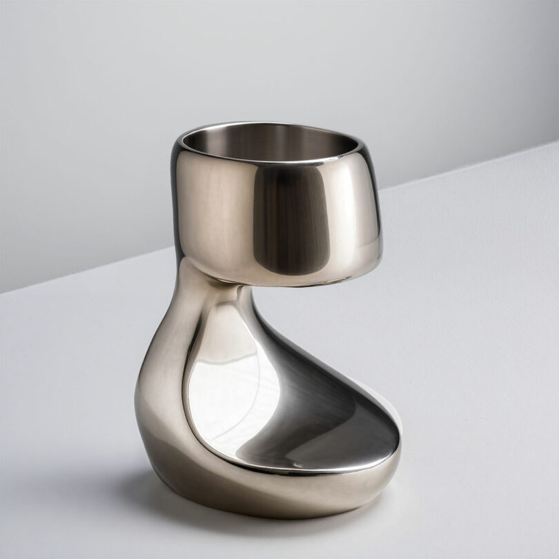 A polished, metallic, modern sculptural cup by Paddy Pike with a thick, curved base sits on a white surface against a light gray background.
