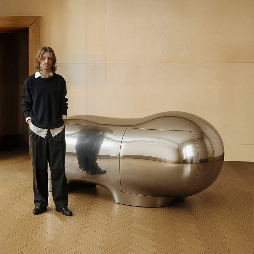A person stands indoors next to a large, metallic, peanut-shaped sculpture by Paddy Pike on a parquet floor against a plain wall.