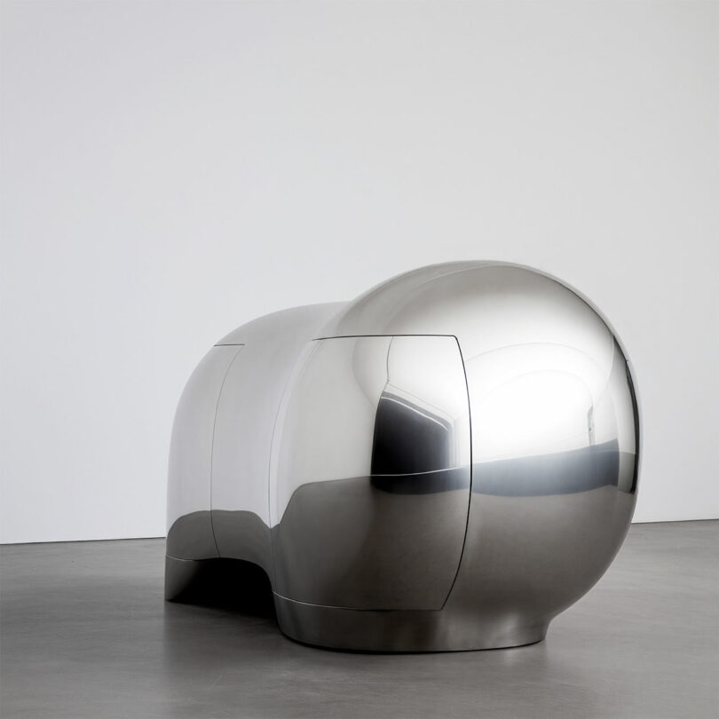 A modern, reflective, silver sculptural object by Paddy Pike, with smooth, rounded shapes, is displayed on a gray floor against a plain white wall.