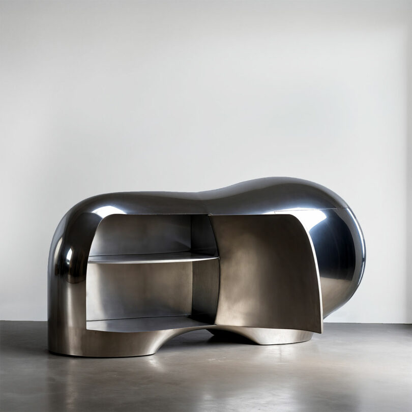 Shiny, metallic, abstract-shaped cabinet by Paddy Pike with curved surfaces, open shelves, and a partially open door, placed on a plain concrete floor against a white wall.