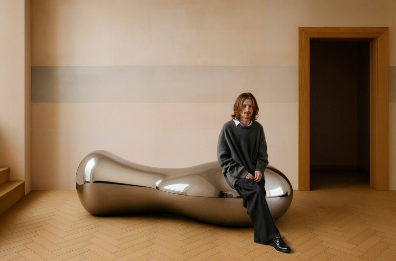 A person with shoulder-length hair sits on a shiny, bean-shaped metallic bench in a minimalist room with beige walls and wooden flooring, echoing the sleek design sensibility of Paddy Pike.
