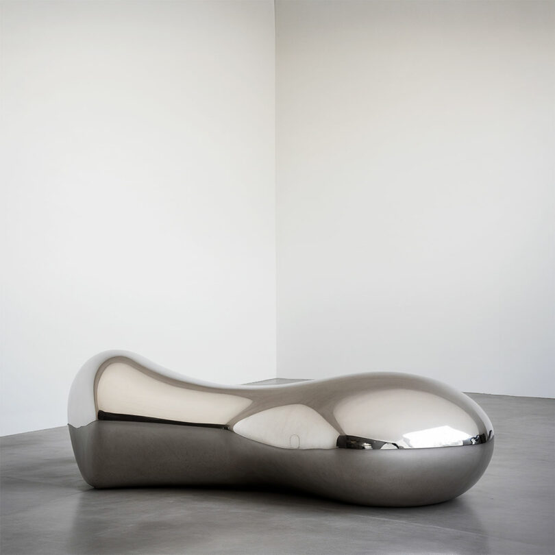 A large, smooth, metallic, bean-shaped sculpture by Paddy Pike sits on a gray concrete floor in a minimal white-walled gallery space.