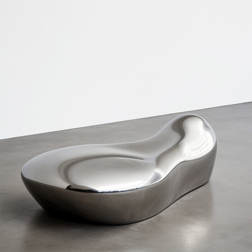 A smooth, metallic, curved bench by Paddy Pike sits on a gray concrete floor against a white wall.