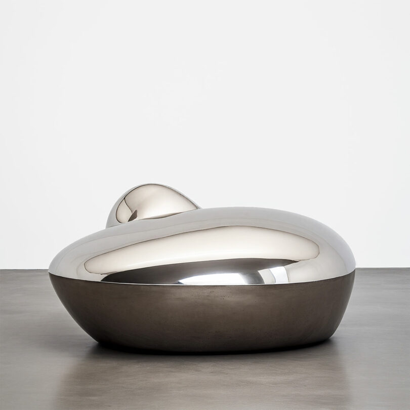A smooth, polished metallic sculpture by Paddy Pike, with rounded, abstract forms, sits on a gray floor against a plain white wall.