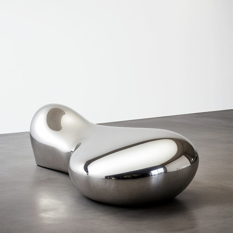 A large, smooth, silver metallic sculpture by Paddy Pike with abstract, rounded shapes sits on a gray floor in a minimalist, white-walled room.