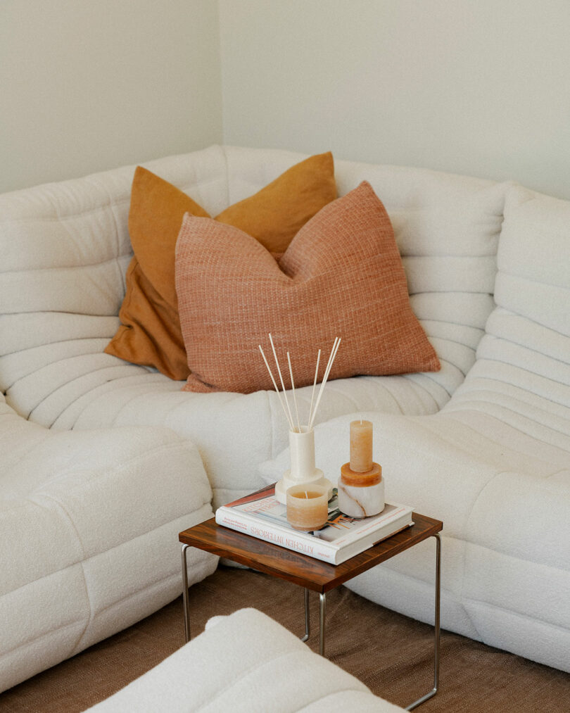 Un sofá seccional blanco con dos cojines naranjas se encuentra junto a una pequeña mesa de madera con velas Stan Editions, un difusor de caña y libros apilados.