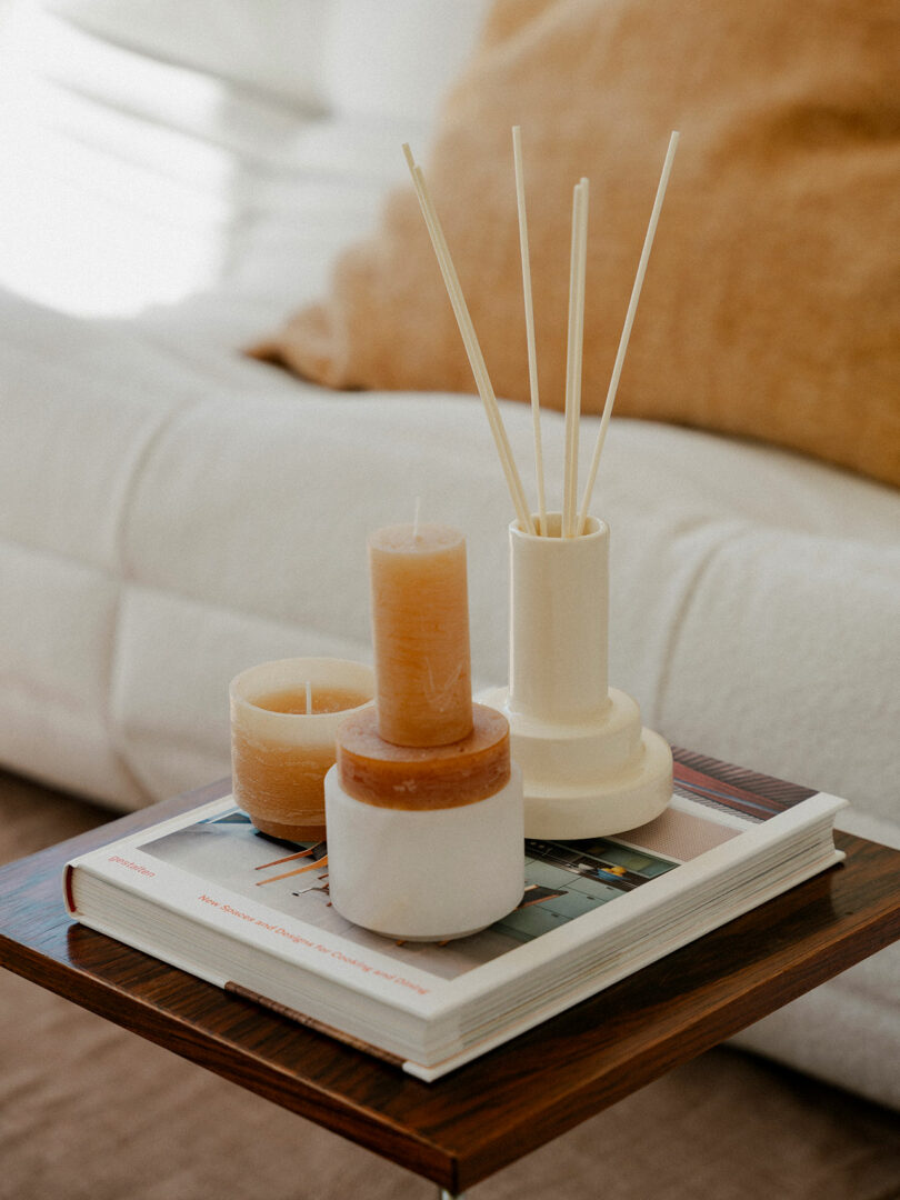 Tres velas Stan y un difusor de caña descansan sobre un libro sobre una mesa auxiliar de madera contra un sofá blanco y almohadas color canela al fondo.