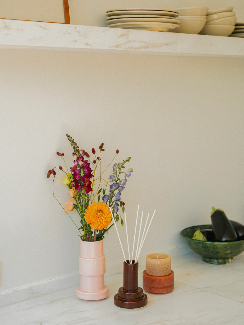 La encimera de mármol contiene un jarrón rosa con flores frescas, un difusor de caña Stan Brown, velas apiladas y un cuenco verde de berenjenas con cuencos apilados debajo del estante.